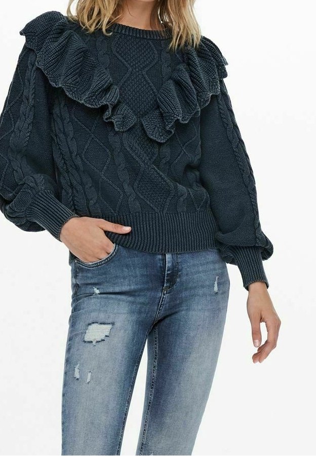 Femme portant un pull torsadé bleu foncé avec des détails volantés et un jean bleu clair délavé, main dans la poche, sur fond blanc.