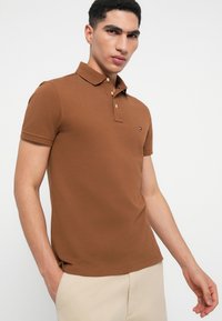 Jauns vīrietis, ģērbies brūnā īsām piedurknēm polo krekla un bēšās biksēs, stāv uz vienkārša balta fona.