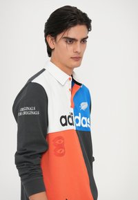 Barvno razdeljen Adidas dolgi rokav polo s belim ovratnikom, ki vključuje modre, oranžne in črne odseke ter opazne logotipe in grafične podrobnosti.