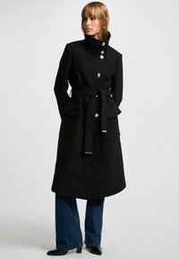 Abrigo de lana negro con cuello alto, botones dorados, cintura ceñida y dos bolsillos frontales, combinado con vaqueros de pierna ancha azules y botas negras hasta el tobillo.