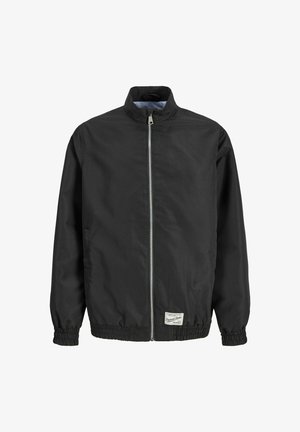 Jongen met krullend haar, gekleed in een zwart jasje met rits, geruite korte broek, witte sokken en zwarte schoenen, staand tegen een witte achtergrond.