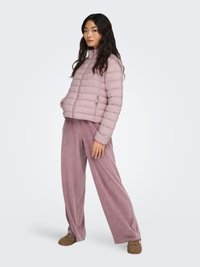 Doudoune rose clair avec coutures horizontales, col montant et fermeture éclair ; associée à un pantalon ample rose pâle doux et des chaussures marron.