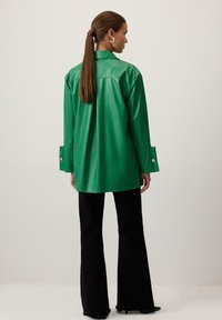 Zaļš, spīdīgs mākslīgās ādas krekla žakets ar brīvu piegriezumu, lielu apkakli un pogāmiem piedurkņiem, kombinēts ar melniem platiem biksēm.