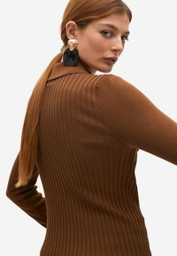 Suéter marrón con diseño de cuello. Presenta una tela texturizada y una silueta ajustada, combinado con grandes pendientes llamativos.