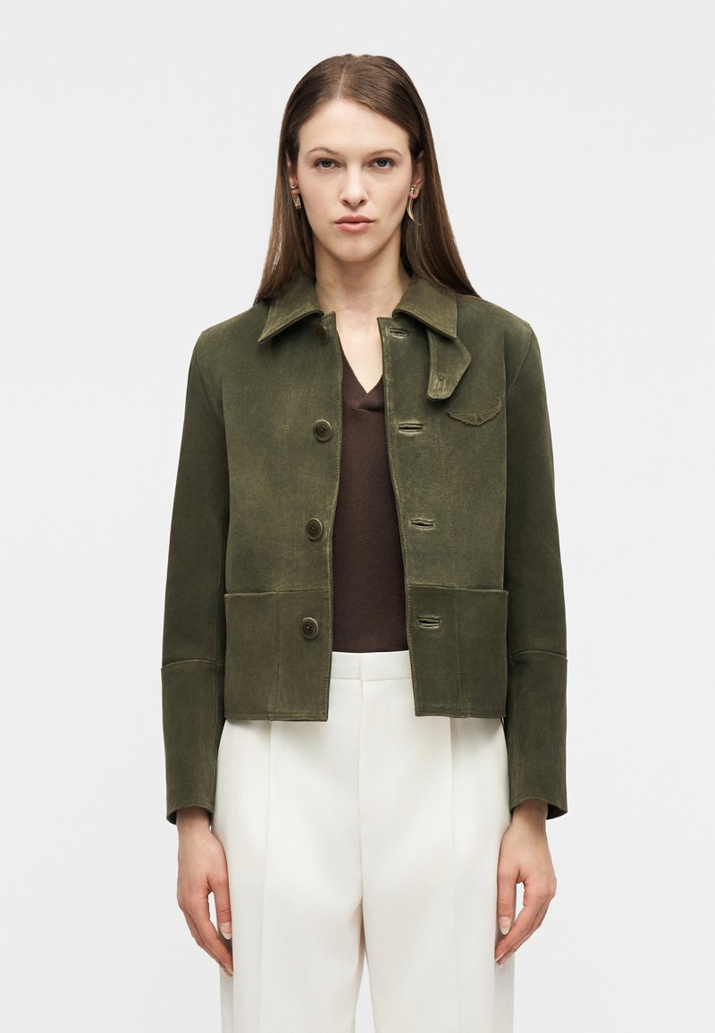 Γυναίκα με σουέντ μπουφάν σε ελιά πράσινο χρώμα πάνω από καφέ τοπ και λευκό παντελόνι με ψηλή μέση, στέκεται μπροστά σε απλό λευκό φόντο.