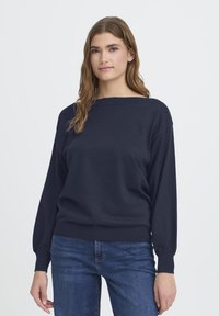 Hosszú barna hajú nő, aki sötétkék, hosszú ujjú pulóvert és kék farmernadrágot visel, egy sima fehér háttér előtt áll.