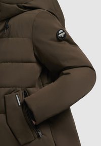 Bruine pufferjack met een gladde textuur, voorzien van een zwart logo-patch op de mouw en een met rits sluitende zak op de borst.
