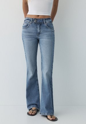 Osoba oblečená v svetlomodrých džínsoch s vysokým pásom a bootcut strihom, bielom krátkom tielku a čiernych sandáloch typu thong, stojaca pred jednoduchým pozadím.