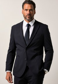 Completo blu navy con giacca a due bottoni, rever a punta e due tasche frontali. Camicia bianca sottostante, abbinata a una cravatta scura. Tessuto dalla texture liscia.