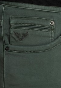 Los vaqueros de mezclilla verdes presentan un bolsillo trasero con cremallera y cierre de botón, además de un logo con alas en relieve. Tienen una textura suave y un detallado de costura.