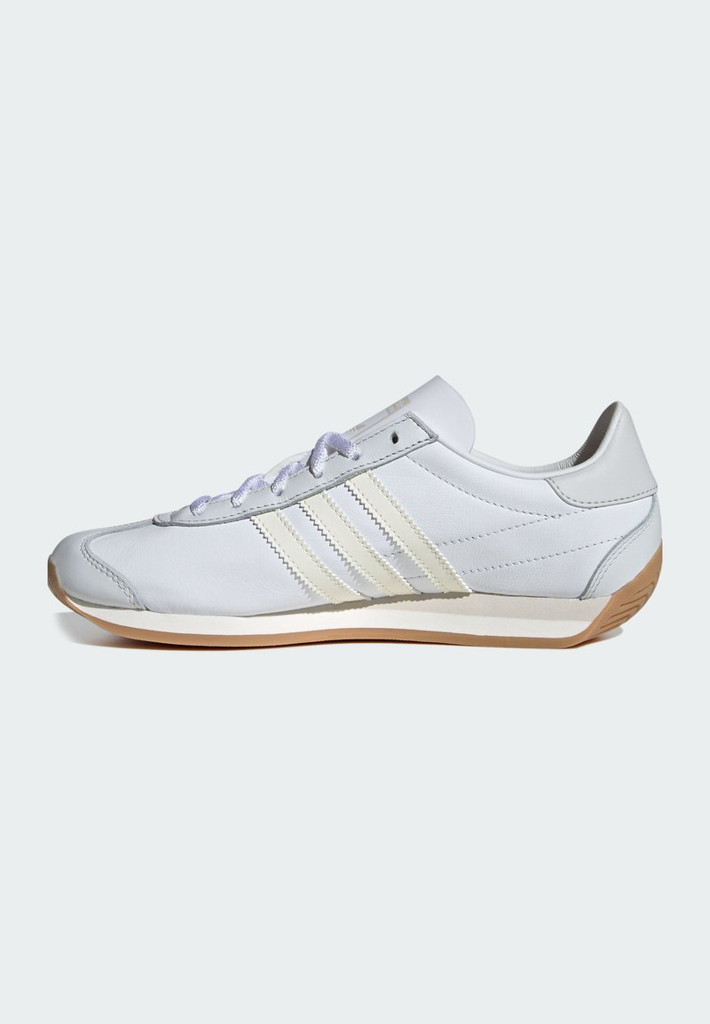 Zapatilla deportiva de cuero blanco con tres franjas crema, punta redondeada y detalles en gris claro. Diseño esbelto y suela de goma texturizada.