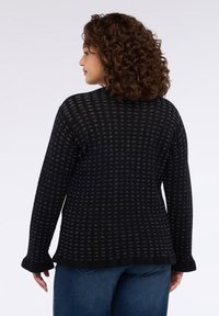 Černý pletený svetr s otevřeným vzorem tkaní, s dlouhými rukávy a žebrovanými manžetami, v kombinaci s modrými džínami.