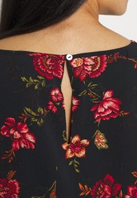 Blus med blommönster som har en svart bakgrund, röda och orange blommor, en nyckelhålsöppning i ryggen och en liten vit knappstängning.