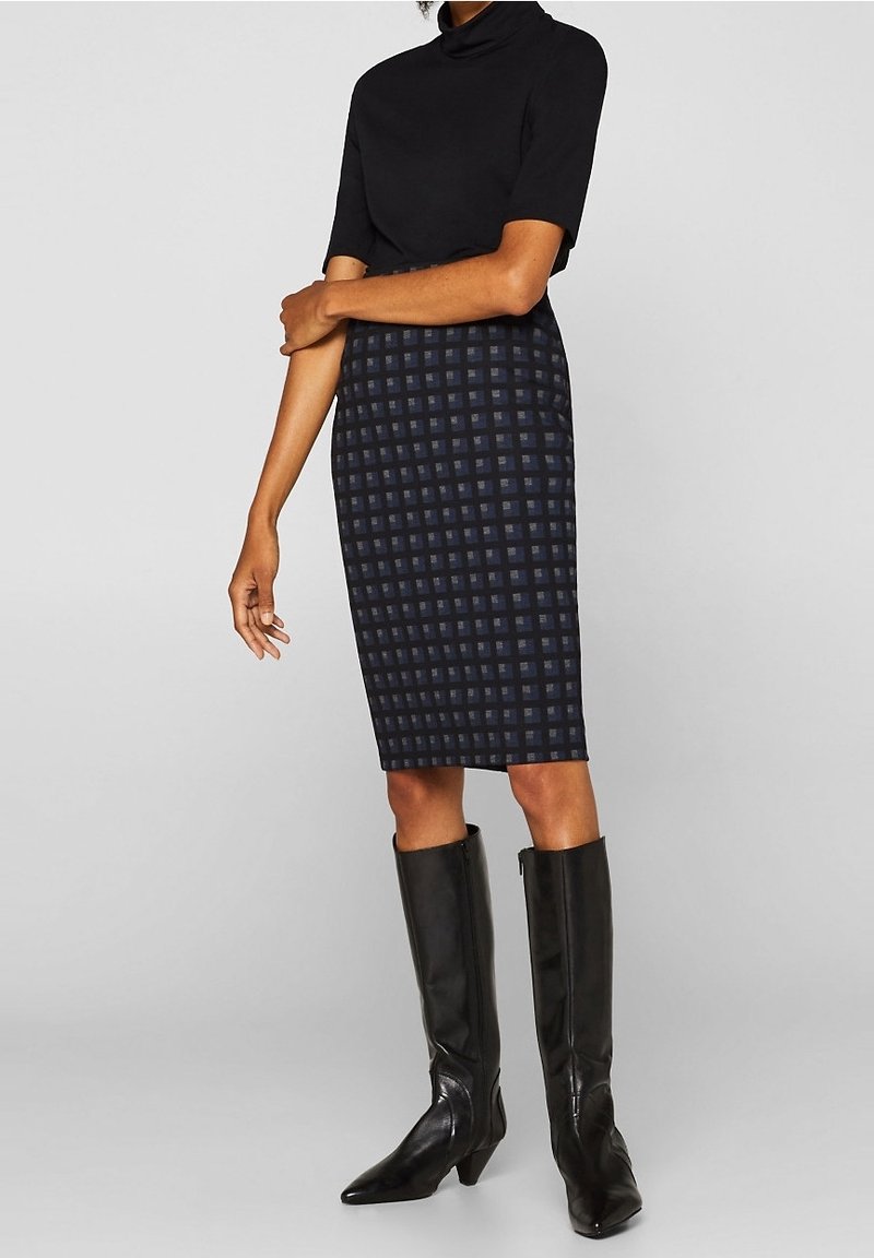Femme portant un col roulé noir à manches courtes, une jupe à carreaux foncés jusqu'aux genoux et des bottes noires montantes, debout devant un fond uni.