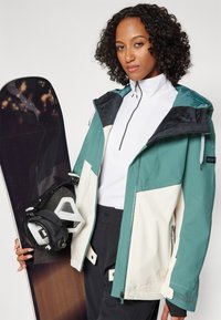 Teal- och cremefärgad färgblock snowboardjacka med avtagbar huva, dragkedja fram och mjuk textur, ihopparad med en svart snowboard och bindningar.
