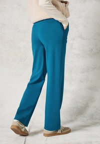 Teal stretchbroek met zijzakken, gekenmerkt door een gladde textuur en een relaxte pasvorm, gecombineerd met lichtgekleurde sneakers tegen een neutrale achtergrond.