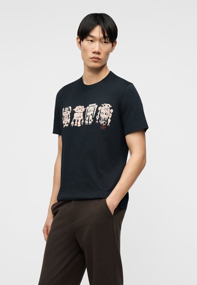 Νέος άντρας που φοράει μαύρο μπλουζάκι με τέσσερα σχέδια ρομπότ σε ανοιχτό μπεζ χρώμα, στέκεται με το ένα χέρι στην τσέπη μπροστά σε απλό ανοιχτό φόντο.