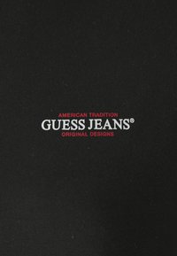 Juodas audinys su tekstūru, ant kurio užrašyta "GUESS JEANS" baltomis raidėmis ir "AMERICAN TRADITION" raudonomis raidėmis viršuje, visiškai centruota.