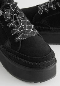 Zapatillas de plataforma de ante negro con cordones texturizados en negro contrastantes, que cuentan con costuras decorativas a lo largo de los bordes y una suela de goma gruesa.