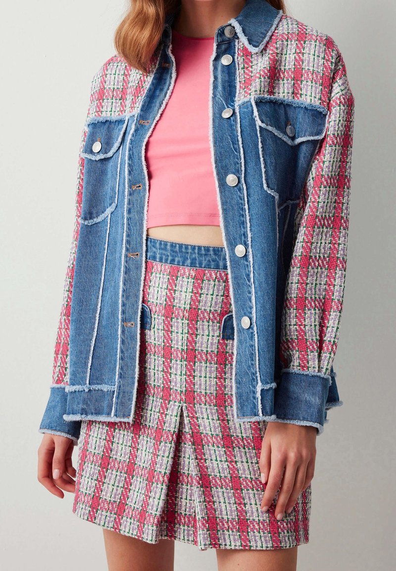 Femme portant un crop top rose, une jupe à carreaux et une veste à carreaux assortie avec des panneaux en denim et des boutons argentés.