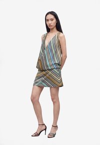 Femme debout portant une robe sans manches à col en V avec des rayures verticales et diagonales multicolores, assortie à des sandales noires à talons hauts à brides.