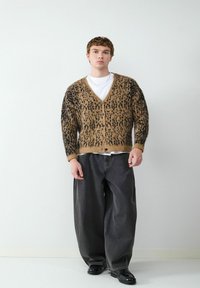 Bruine en zwarte cardigan met een luipaardpatroon, met een V-hals en knopen, gedragen over een witte T-shirt, gecombineerd met losse, donkere jeans.