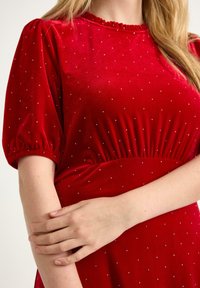 Czerwona sukienka z aksamitnego materiału, z krótkimi bufkami, marszczoną talią i drobnymi srebrnymi akcentami. Teksturowany materiał z delikatnym połyskiem, okrągły dekolt.