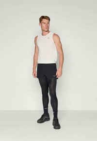 Uomo in forma che indossa una maglietta senza maniche bianca, pantaloni sportivi neri sopra leggings neri, calze nere e scarpe da corsa nere, in piedi e che guarda a destra.
