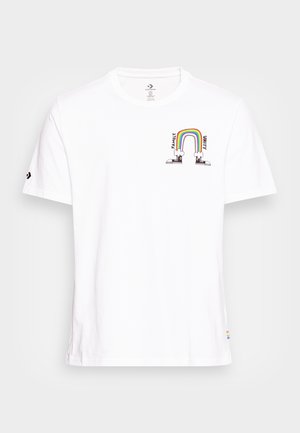 T-shirt en coton blanc avec un col rond. Présente un graphique imprimé arc-en-ciel au-dessus du texte "FAMILLE" et "UNITE" avec deux chaussures de dessin animé en bas.