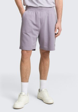 Mann trägt hellviolette Shorts mit elastischem Bund, weiße Kniestrümpfe und weiße Turnschuhe, steht mit einer Hand in der Tasche.
