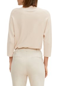 Maglione beige lavorato a maglia con colletto alto, maniche a tre quarti e spalle texturizzate, abbinato a pantaloni beige chiaro con taglio sartoriale e tasche posteriori.
