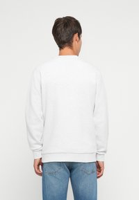 Homme de dos portant un sweat-shirt gris clair uni et un jean en denim bleu devant un arrière-plan uni.