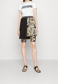 Vit T-shirt med svart text "Versace Jeans Couture", kombinerad med en svart och guldmönstrad kjol och svarta sandaler. Har veck och grafiska tryck.