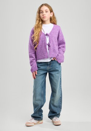 Paarse gebreide cardigan met twee voorknoopjes, gedragen over een wit shirt, gecombineerd met losse blauwe jeans en lichte sneakers.