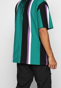 T-shirt w paski w kolorach: turkusowym, czarnym i fioletowym. Krótkie rękawy, luźny krój oraz widoczna szew na plecach. Materiał bawełniany.