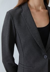 Blazer gris con acabado texturizado, solapas de pico y un botón negro único, ajuste entallado con un bolsillo frontal. Camiseta negra sin mangas debajo.