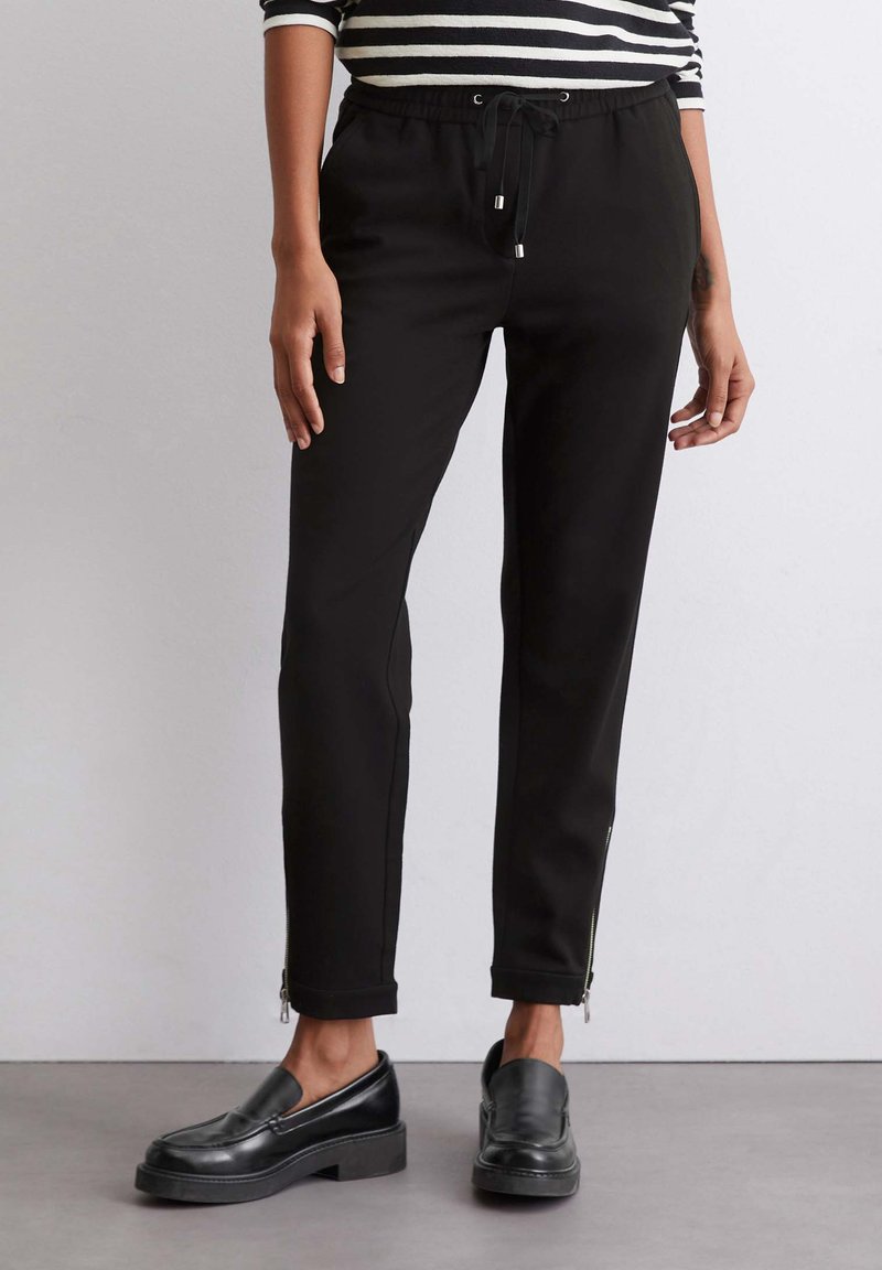 Zwarte joggingbroek met elastische tailleband en trekkoord, met ritsdetails bij de boorden en een gladde, gestructureerde stof.
