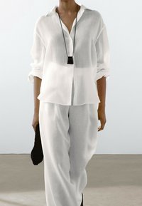 Camicia bianca leggera realizzata in tessuto trasparente, con bottoni e maniche arrotolate; abbinata a pantaloni larghi coordinati e una pochette nera.