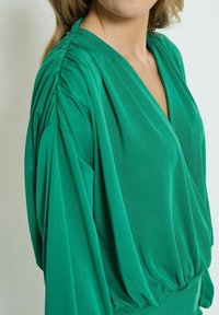 Top in tessuto verde con scollatura a V, dettagli arricciati sulle spalle e fondo morbido con elastico. Texture liscia e design drappeggiato.