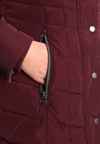 Main insérée dans la poche zippée d'une veste matelassée bordeaux avec des boutons-pression et un logo visible sur la tirette du zip.
