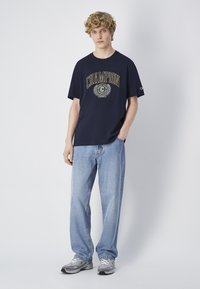 Camiseta navy con gráfico "Champion" en dorado y diseño de laurel, combinada con jeans de color azul claro y zapatillas grises. Estilo y ajuste casual.