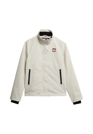 Chaquetón beige claro con un cuello alto, bolsillos con cremallera y detalles en negro en los puños; presenta un parche con el logotipo en la parte superior izquierda del pecho.