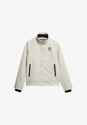 Chaquetón beige claro con un cuello alto, bolsillos con cremallera y detalles en negro en los puños; presenta un parche con el logotipo en la parte superior izquierda del pecho.