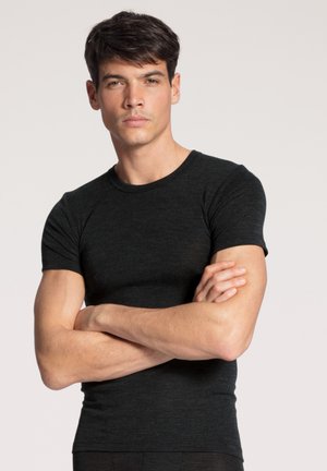 Jonge man met kort donker haar, draagt een aansluitend zwart T-shirt, armen over elkaar, staand tegen een effen lichte achtergrond.
