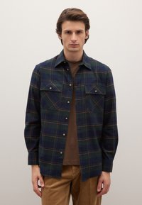 Camisa de franela a cuadros en azul marino y verde con detalles en naranja, con bolsillos con botones y textura suave. Lleva puesta sobre una capa base marrón.