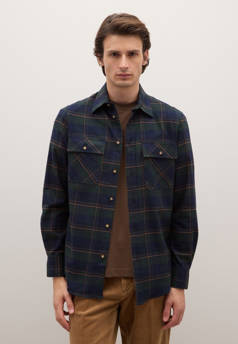 Camisa de franela a cuadros en azul marino y verde con detalles en naranja, con bolsillos con botones y textura suave. Lleva puesta sobre una capa base marrón.