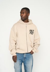 Sudadera con cremallera color beige en tejido suave con bolsillo frontal. Presenta un estampado gráfico en el lado izquierdo del pecho y capucha integrada.
