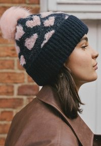 Young woman in profile wearing a black and pink fuzzy knit hat with pom-pom and a brown leather jacket against a brick wall background.