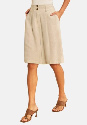 Short beige mi-long aux genoux, jambes larges, avec boutons et poches à l’avant, porté avec des sandales à talons ouvertes marron.