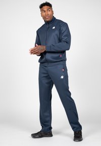 Marine blauwe trainingspak van soepel materiaal met een hoge kraag. Bevat een logo op de borst, zijzakken en contrasterende accenten op de broek.
